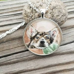 May include: A silver chain necklace with a round glass pendant featuring a watercolor painting of a dog's face. The dog has brown and black fur and is looking directly at the viewer.