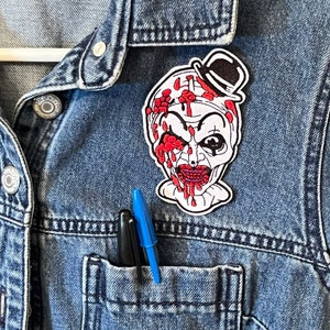 May include: A white embroidered patch of a clown with a red and white hat, dripping with blood, on a blue denim jacket.