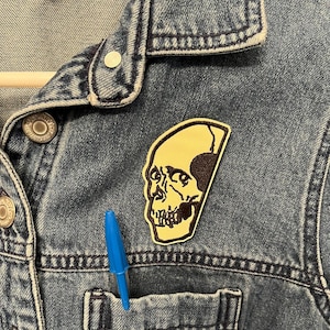 May include: A brown skull patch sewn onto a blue denim jacket. The skull is outlined in yellow and has a detailed design.