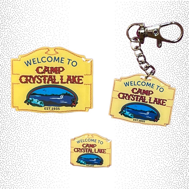 Puede incluir: Se&ntilde;al rectangular amarilla de Camp Crystal Lake con el texto "WELCOME TO CAMP CRYSTAL LAKE" y "EST.1935". Una se&ntilde;al es un llavero y dos son independientes. La se&ntilde;al presenta una escena de lago.