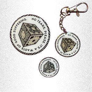May include: A black and white keychain with a circular metal charm featuring a cube with the text "IT'S A WASTE OF GOOD SUFFERING. NO TEARS, PLEASE."