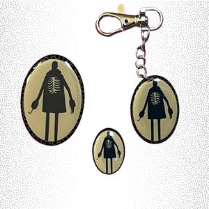 May include: A set of three keychains featuring a black silhouette of a person with a skeleton inside, wearing a coat. The keychains are made of resin and have a gold-colored metal ring.
