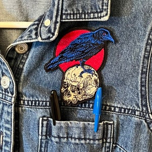 May include: A blue denim jacket with a red and blue embroidered patch of a crow perched on a skull. Two pens are in the pocket of the jacket.