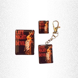 May include: Three black and red enamel pins with the text "Late Night with the Devil" and a graphic of a person with flames on their head. The pins are attached to a gold key chain.