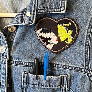 May include: A denim jacket with a heart-shaped patch featuring a black and white illustration of Frankenstein's monster and his bride kissing. The patch has a jagged edge and is sewn onto the jacket's left chest pocket. Two pens are sticking out of the pocket.