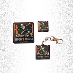 May include: Three different sizes of a black and orange enamel pin featuring a skeleton dancing with the text "Night Owls" and the artist's signature.