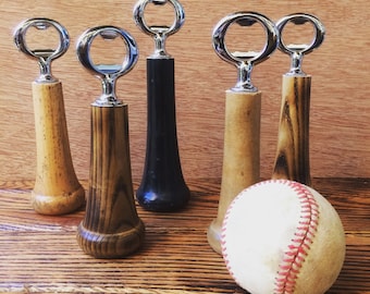 May include: A collection of bottle openers with wooden and black handles, and a baseball. The openers have a silver metal top. The wooden handles vary in color and grain. The baseball is white with red stitching.