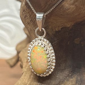 May include: An oval-shaped cabochon opal pendant set in a sterling silver bezel with a decorative beaded and filigree edge. The opal displays flashes of orange, yellow, and green fire. The pendant hangs from a thin sterling silver box chain.