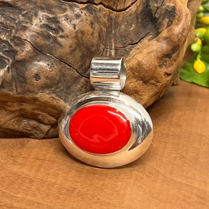 May include: A round, silver pendant with a vibrant red center. The pendant has a polished finish and a loop at the top for a chain. The red center is smooth and glossy, creating a striking contrast with the silver.