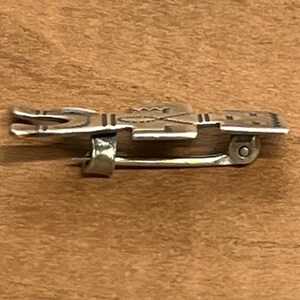 May include: A silver-toned metal brooch with a detailed design. The brooch features a rectangular shape with etched patterns and a pin closure on the back. The brooch is displayed on a wooden surface.