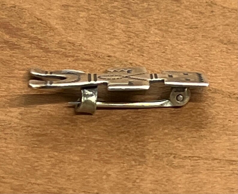 May include: A silver-toned metal brooch with a detailed design. The brooch features a rectangular shape with etched patterns and a pin closure on the back. The brooch is displayed on a wooden surface.