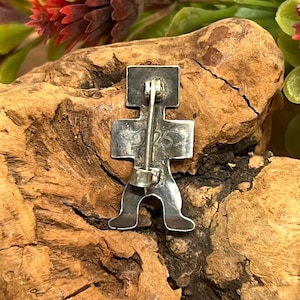May include: A silver-colored pin in the shape of a stylized figure. The pin has a rectangular head, a cross-shaped body, and two feet. The pin is attached to a silver bar with a clasp on the back. The pin is set against a natural wood background.