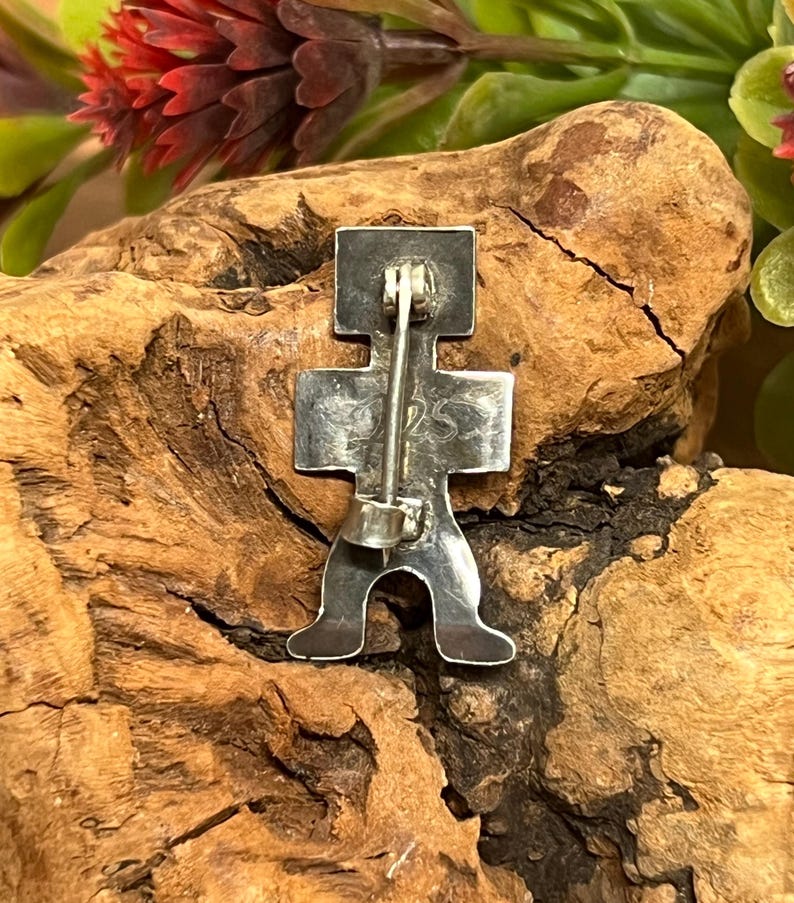May include: A silver-colored pin in the shape of a stylized figure. The pin has a rectangular head, a cross-shaped body, and two feet. The pin is attached to a silver bar with a clasp on the back. The pin is set against a natural wood background.