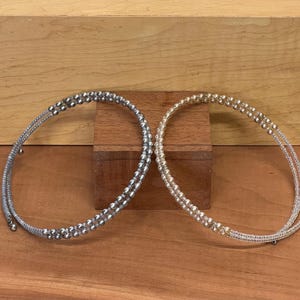 May include: Two beaded chokers. One is silver, the other is gold. Each choker is made of small, round beads strung on a wire. The chokers are displayed on a wooden surface.