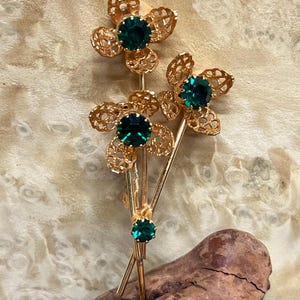 May include: A gold-toned floral brooch with three flower clusters. Each flower has a dark green gemstone center and openwork petals. The brooch is set against a light-colored, textured background.