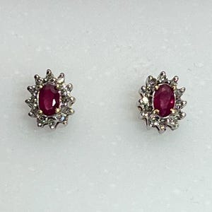 Puede incluir: Un par de pendientes de botón con piedras rojas ovaladas, rodeadas por un halo de piedras claras más pequeñas. Los pendientes tienen un engaste de metal plateado, creando un aspecto clásico y elegante. Las piedras miden aproximadamente 0,6 cm de largo.