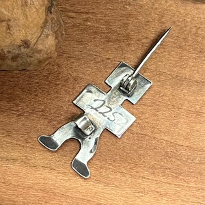 May include: A silver-toned metal pin in the shape of a stick figure. The pin has a long, sharp needle and a clasp on the back. The number "225" is stamped on the figure. The pin is on a wooden surface.