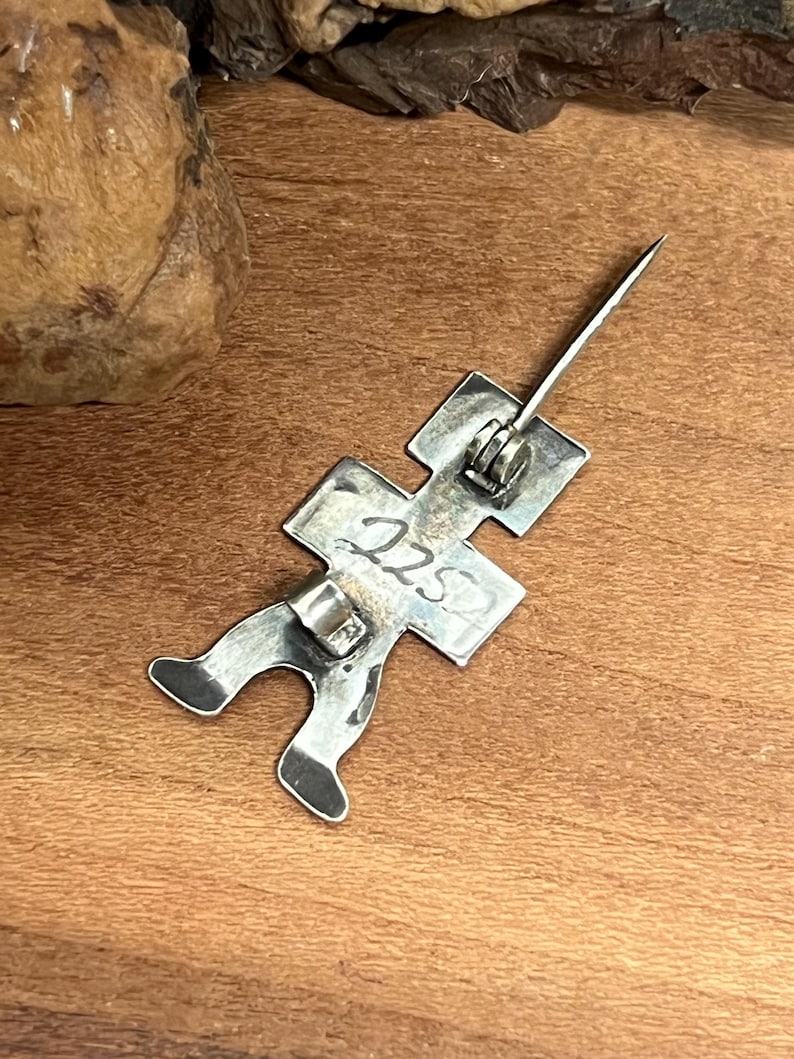 May include: A silver-toned metal pin in the shape of a stick figure. The pin has a long, sharp needle and a clasp on the back. The number "225" is stamped on the figure. The pin is on a wooden surface.