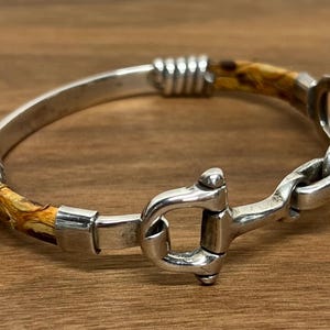 以下が含まれることがあります： 磨き上げられた金属製のビット型クラスプと、茶色とベージュの模様が入った質感のある素材の2つの湾曲したセグメントを備えたシルバーブレスレット。デザインには、滑らかなシルバーのエンドキャップと、金属と素材の接合部に巻かれたワイヤーのアクセントが含まれています。