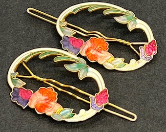 May include: Two oval-shaped hair clips featuring gold-tone metal frames with enamel detailing. The design includes colorful floral motifs in shades of orange, purple, and pink, accented by green leaves and white enamel borders on the oval structures.