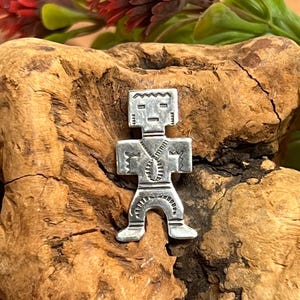 May include: A silver-colored pendant in the shape of a stylized human figure. The pendant has a rectangular head with etched details, a zipper-like design down the torso, and outstretched arms. The pendant is displayed on a piece of wood.