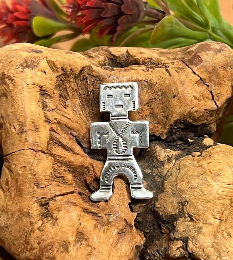 May include: A silver-colored pendant in the shape of a stylized human figure. The pendant has a rectangular head with etched details, a zipper-like design down the torso, and outstretched arms. The pendant is displayed on a piece of wood.