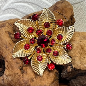 Könnte beinhalten: Eine goldfarbene Blumenbrosche mit roten Edelsteinakzenten. Die Brosche hat einen zentralen großen roten Edelstein, der von kleineren roten Edelsteinen und goldfarbenen blattförmigen Elementen umgeben ist. Die Brosche ist vor einem hölzernen Hintergrund platziert.
