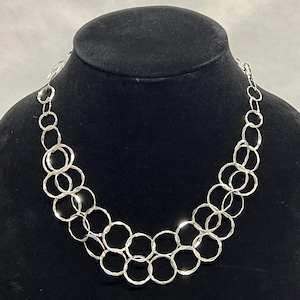 Puede incluir: Un collar plateado con una serie de anillos texturizados interconectados. El collar se exhibe en un maniquí negro, mostrando su diseño en capas y superficie reflectante. La pieza de joyería es un accesorio llamativo.
