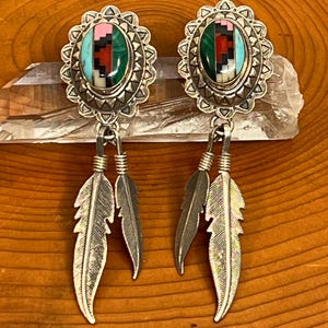 Könnte beinhalten: Silberne Ohrringe mit ovalen, mehrfarbigen Steineinlagen und baumelnden Federanhängern. Die Ohrringe haben ein Design aus Türkis, Rosa, Rot und Grün, das in einen dekorativen Silberrahmen gefasst ist. Die Federanhänger sorgen für Bewegung.