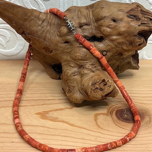 May include: A necklace composed of cylindrical, porous, orange-red beads with a textured surface. The beads are strung together and secured with a small, metallic, cylindrical screw-style clasp. The necklace features variations in color intensity.
