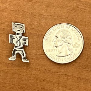 May include: A small, silver-colored pendant in the shape of a stylized human figure, with a quarter coin next to it for size comparison. The pendant has a geometric design and is set against a brown wooden surface.