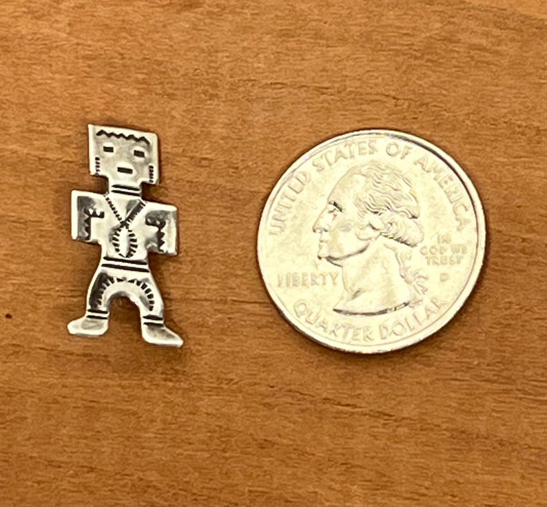 May include: A small, silver-colored pendant in the shape of a stylized human figure, with a quarter coin next to it for size comparison. The pendant has a geometric design and is set against a brown wooden surface.