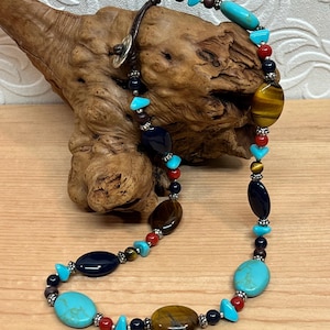 Puede incluir: Un collar con cuentas turquesas, marrones, rojas y azul marino. El collar incluye cuentas ovaladas y redondas, un cordón de cuero y detalles metálicos plateados. El collar se exhibe sobre una superficie de madera.