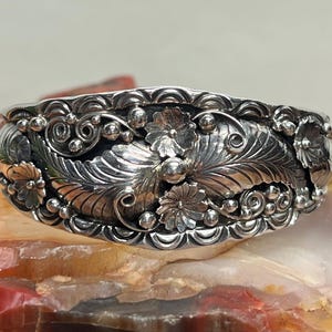 May include: A silver cuff bracelet with intricate floral and leaf designs. The bracelet features detailed carvings of flowers, leaves, and swirling patterns. The metal has a polished finish, highlighting the detailed craftsmanship of the jewelry.