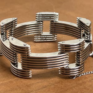 May include: A silver-toned bracelet with a unique interlocking design. The bracelet features multiple curved, layered segments connected to form a chain. The metal has a polished, reflective surface, and a delicate chain clasp.