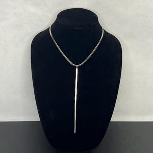 May include: A silver-toned necklace with a sleek, minimalist design. The necklace features a thin, flat chain that forms a V-shape, with a long, rectangular bar pendant suspended from the center. The pendant hangs vertically, creating a modern and elegant look.