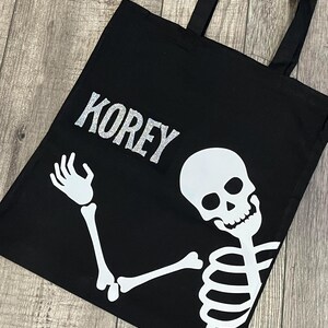 Puede incluir: Bolsa de lona negra con un diseño de esqueleto blanco y el nombre "KOREY" en letras de purpurina plateada.