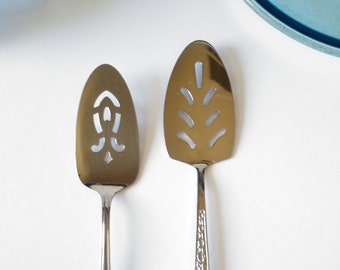 May include: Two silver cake servers with decorative cutouts. The server on the left has a more ornate design with a pointed tip. The server on the right has a more simple design with a rounded tip.
