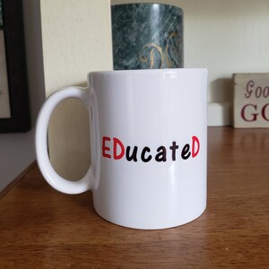May include: White ceramic mug with a black and red "Educated" design. The mug is sitting on a wooden surface.