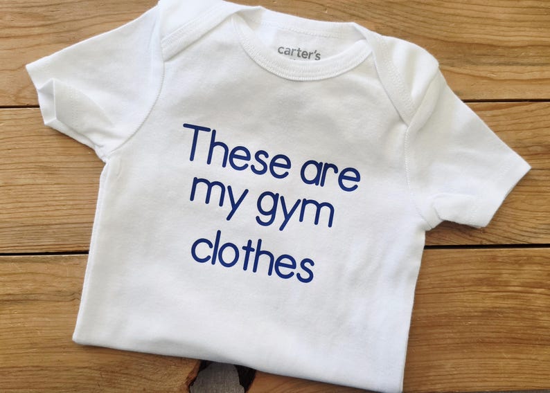 May include: A white short-sleeved baby bodysuit featuring the text "These are my gym clothes" printed in dark blue lettering on the front. The garment has a round neckline and a brand label reading "carter's" visible on the inside collar area.