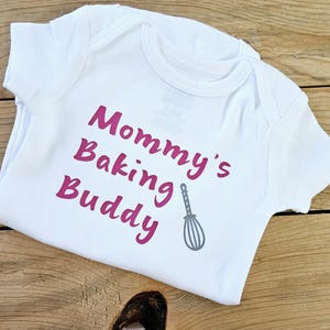 May include: White baby onesie with pink text that reads "Mommy's Baking Buddy" and a gray whisk graphic.