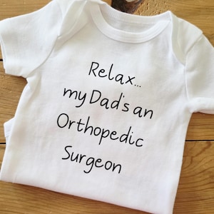 May include: White baby onesie with the text "Relax... my Dad's an Orthopedic Surgeon" in black script. The onesie is made of soft fabric and has short sleeves. The background is a light brown wooden surface.