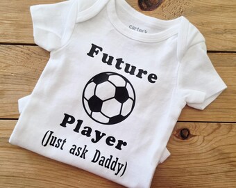 May include: White baby onesie with the text "Future Player (Just ask Daddy)" in black, along with a soccer ball graphic. The onesie is made of soft material and is designed for a baby.