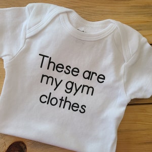 May include: A white short-sleeved baby bodysuit featuring the black printed text "These are my gym clothes" centered on the front. The garment has a round neckline and short sleeves with ribbed trim detailing around the collar and leg openings.