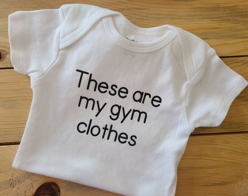 May include: A white short-sleeved baby bodysuit featuring the black printed text "These are my gym clothes" centered on the front. The garment has a round neckline and short sleeves with ribbed trim detailing around the collar and leg openings.