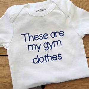 May include: A white short-sleeved bodysuit with a crew neckline and the text "These are my gym clothes" printed in blue on the front. The garment features a "carter's" brand label on the inside of the collar and has a simple, clean design.
