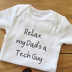 May include: A white short-sleeved baby bodysuit featuring the black printed text "Relax... my Dad's a Tech Guy" on the front. The garment has a round neckline and is labeled with the Carter's brand name on the inside of the collar area.