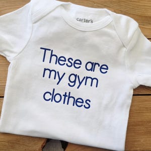 May include: A white short-sleeved baby bodysuit featuring the text "These are my gym clothes" printed in dark blue lettering on the front. The garment has a round neckline and a brand label reading "carter's" visible on the inside collar area.