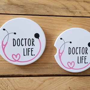 May include: Two white round coasters with a pink stethoscope design and the text "DOCTOR LIFE."