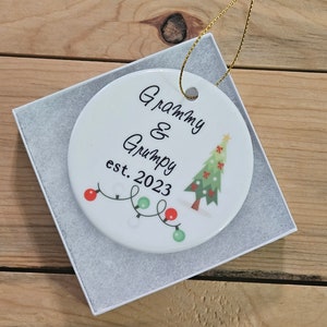 May include: White ceramic Christmas ornament with a green Christmas tree and red and green lights. The ornament says "Grammy & Grumpy est. 2023".